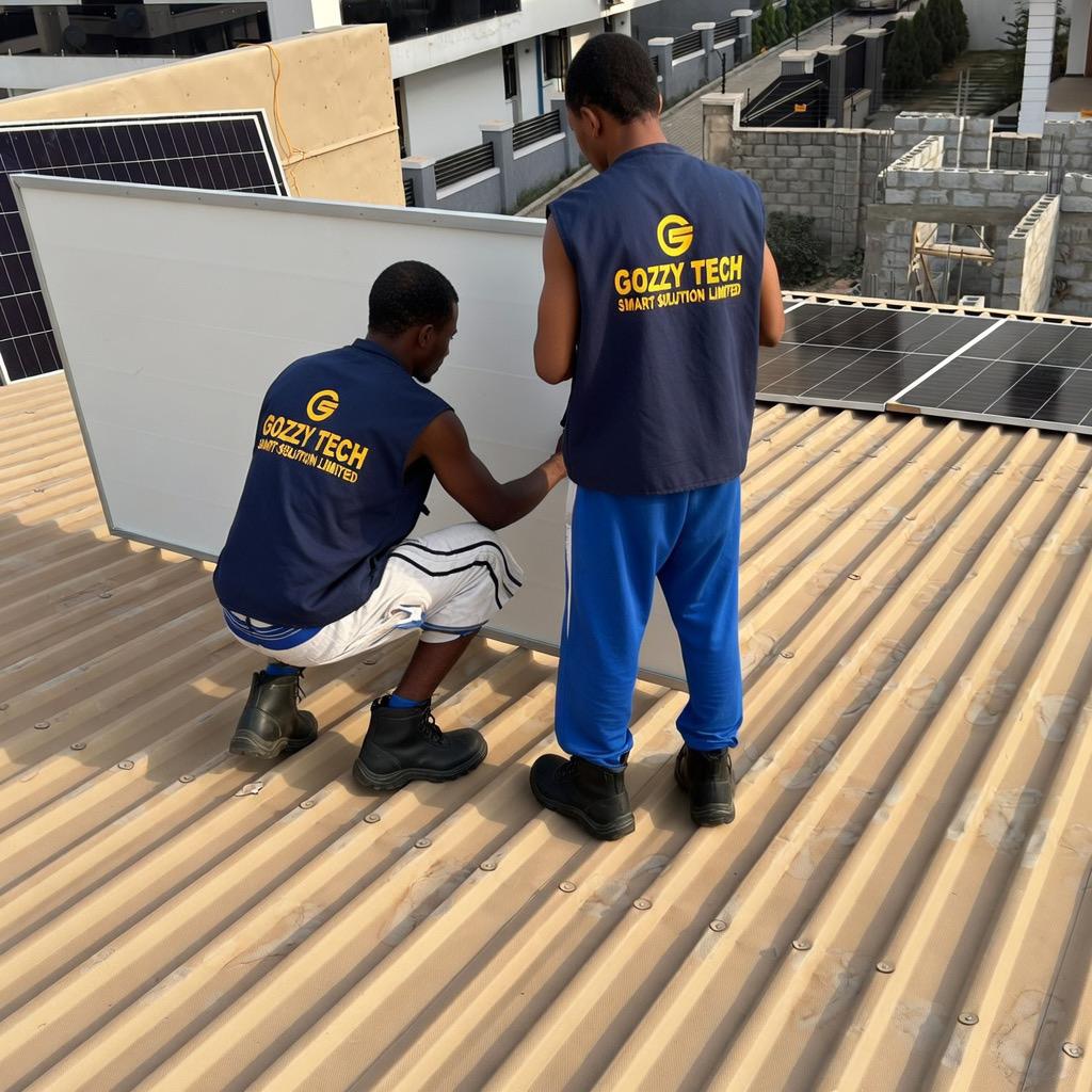 Two GOZZY TECH technicians properly installing a large solar panel on a rooftop in Nigeria. The workers are wearing branded safety vests and safety boots while handling the panel with care on a corrugated roof.